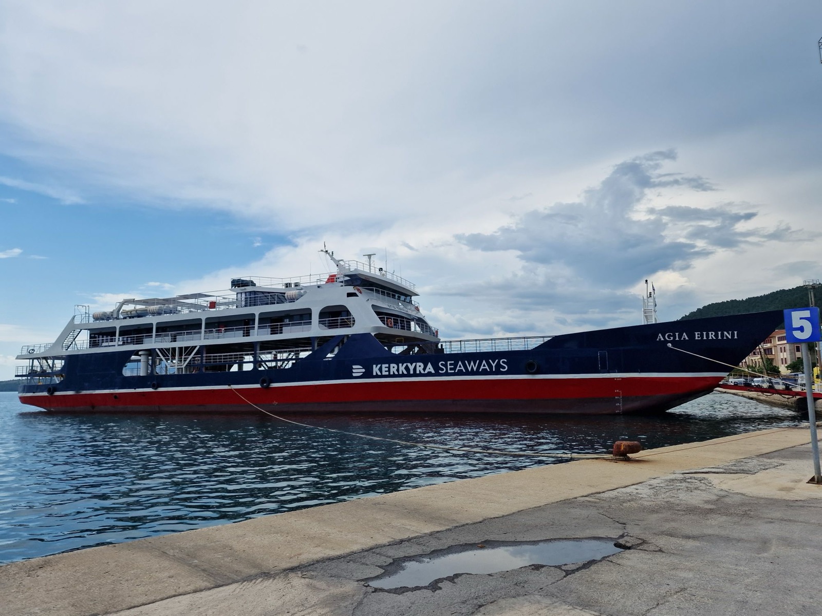 Agia Eirini, a car ferry with a dark navy hull, a red lower hull and the name Kerkyra Seaways in white capitals, an open car deck below a white superstructure, moored at a dock in Igoumenitsa under a cloudy sky.