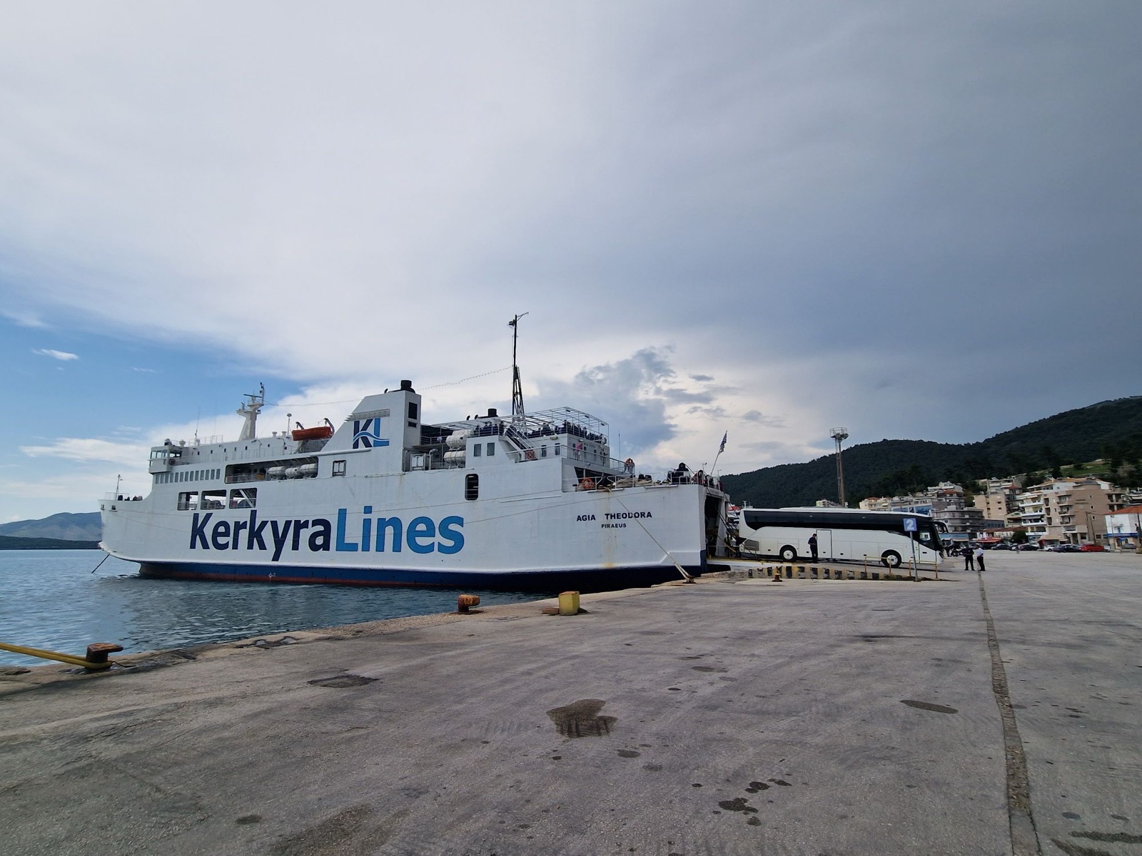 Agia Theodora, a white car ferry with the Kerkyra Lines name in large blue letters and a blue funnel logo, moored at a concrete dock in Igoumenitsa with her stern ramp lowered and a coach boarding, wooded hills and the town behind.