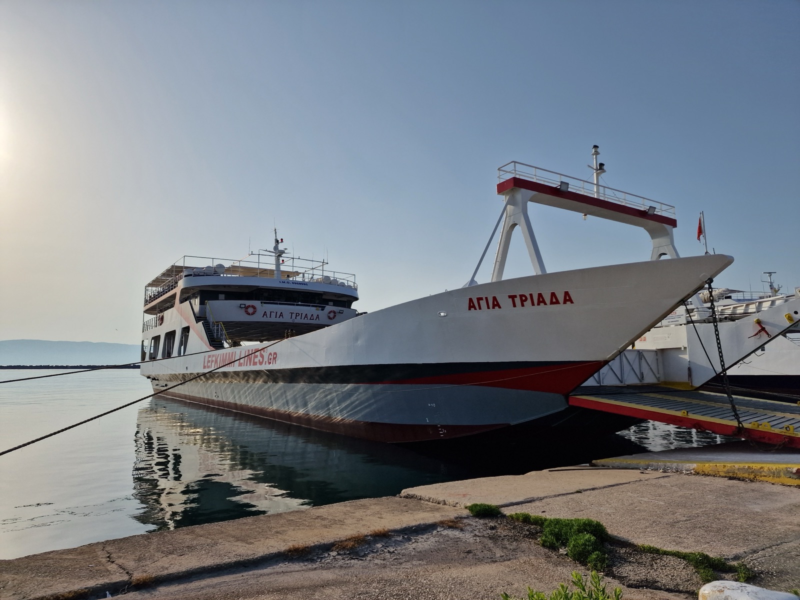Agia Triada, a white car ferry with a red and black stripe above a grey lower hull and the words Lefkimmi Lines.gr along her side, moored at a dock in Lefkimmi, Corfu, with her raised bow ramp and its white gantry at the right, calm water and a pale morning sky.