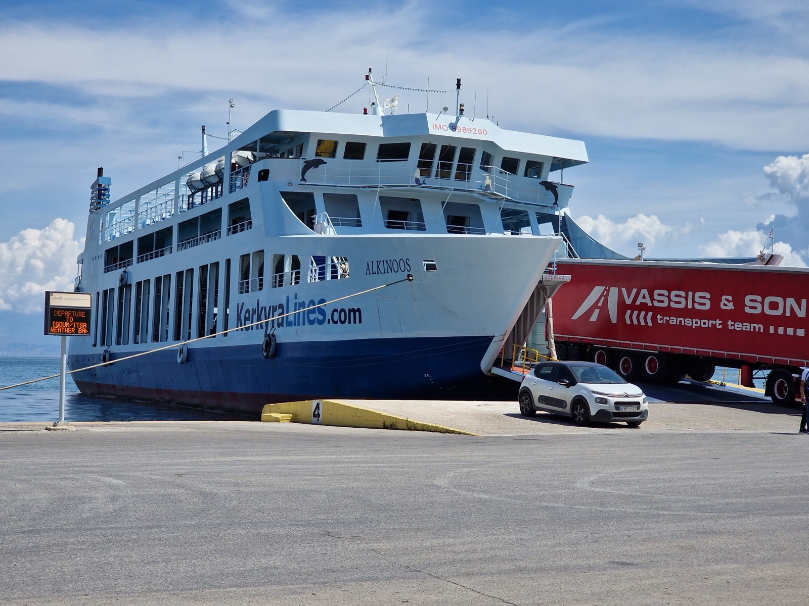 Alkinoos, a white car ferry with a dark blue lower hull and the words KerkyraLines.com along her side, moored bow-first at a dock in Corfu with her bow door open as a car and a red lorry trailer drive aboard, dolphin and mermaid decals on the front of her bridge.