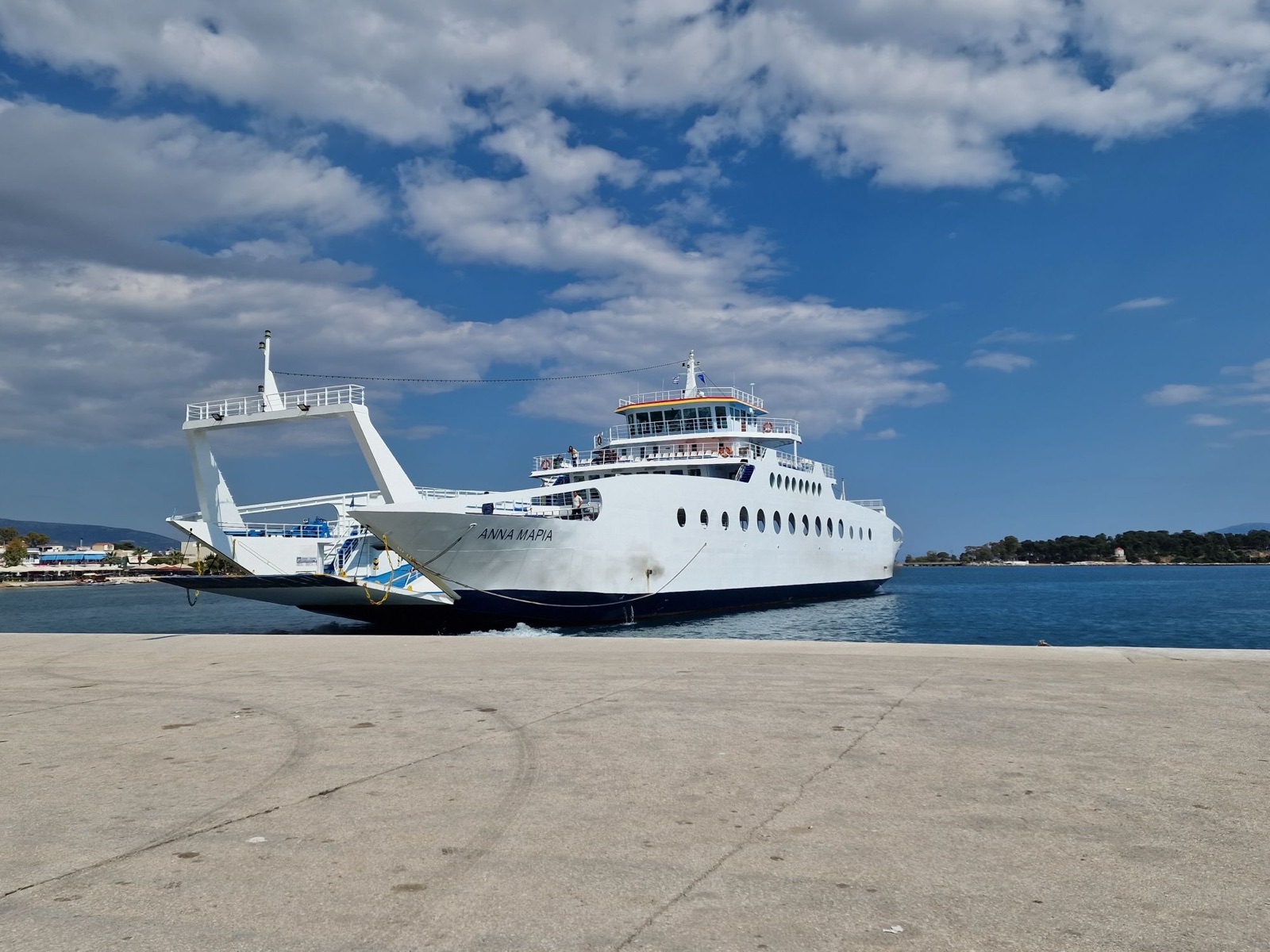 Anna Maria, a white double-ended car ferry with a dark blue lower hull and a row of round portholes, arriving at a concrete dock with her ramp lowered under a blue sky with broken cloud.