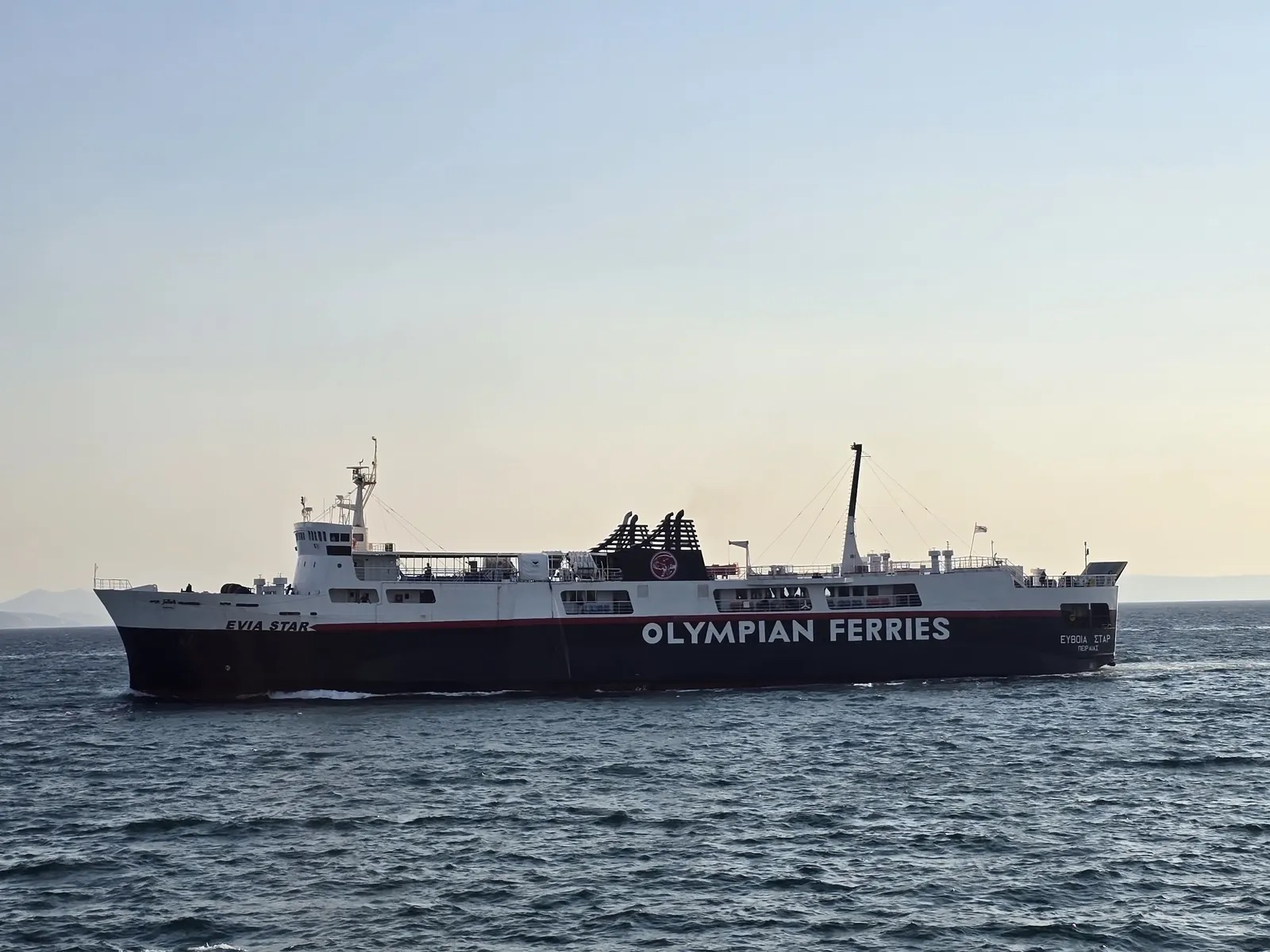 Evia Star under way on a calm sea in Olympian Ferries colours, a ferry with a dark blue hull, red stripe and white superstructure, the company name in large white letters along the side.