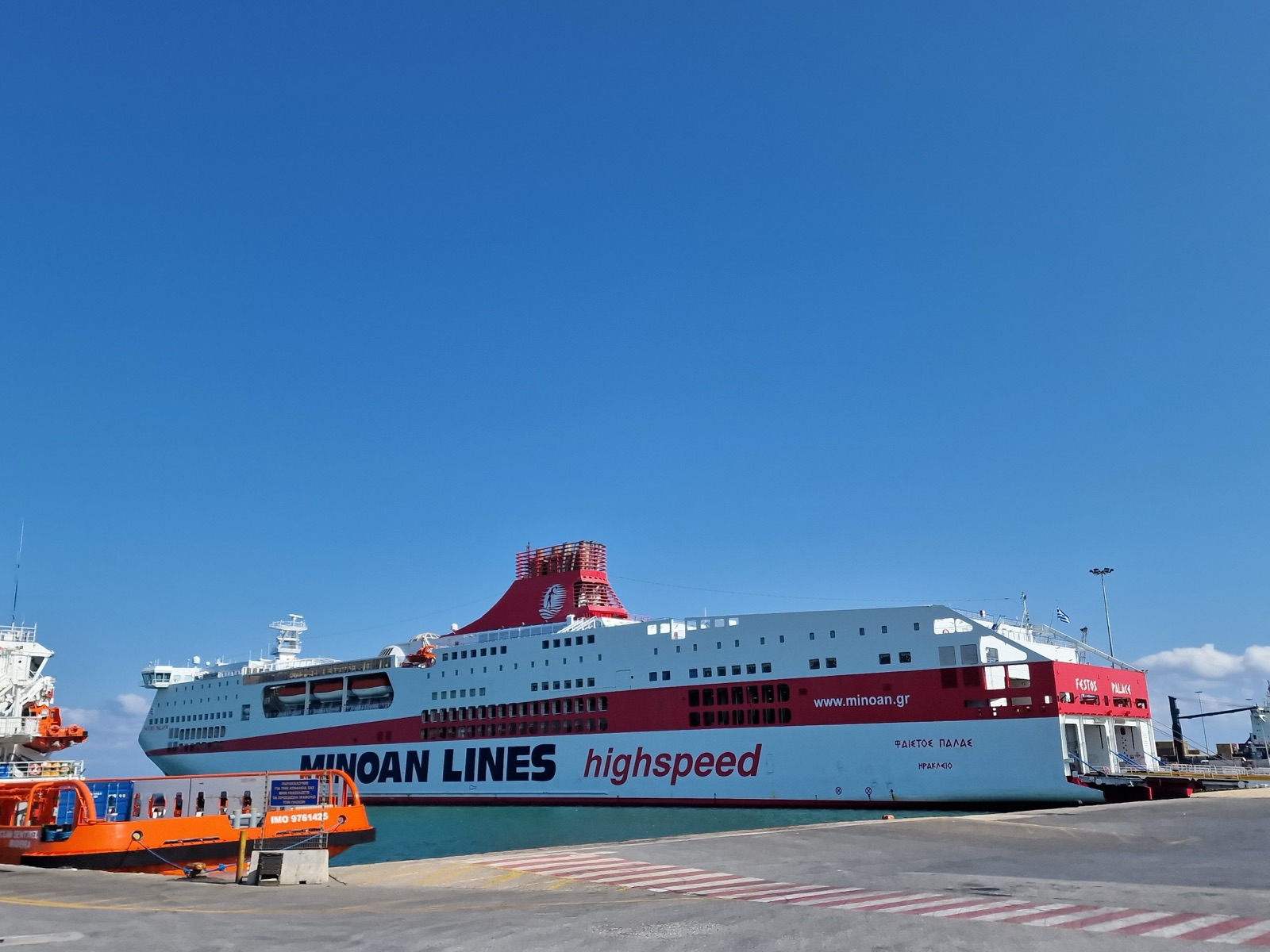 Festos Palace, a large Minoan Lines ferry with a white hull, a broad red band and a red funnel, moored stern-to at the dock in Heraklion under a clear blue sky.