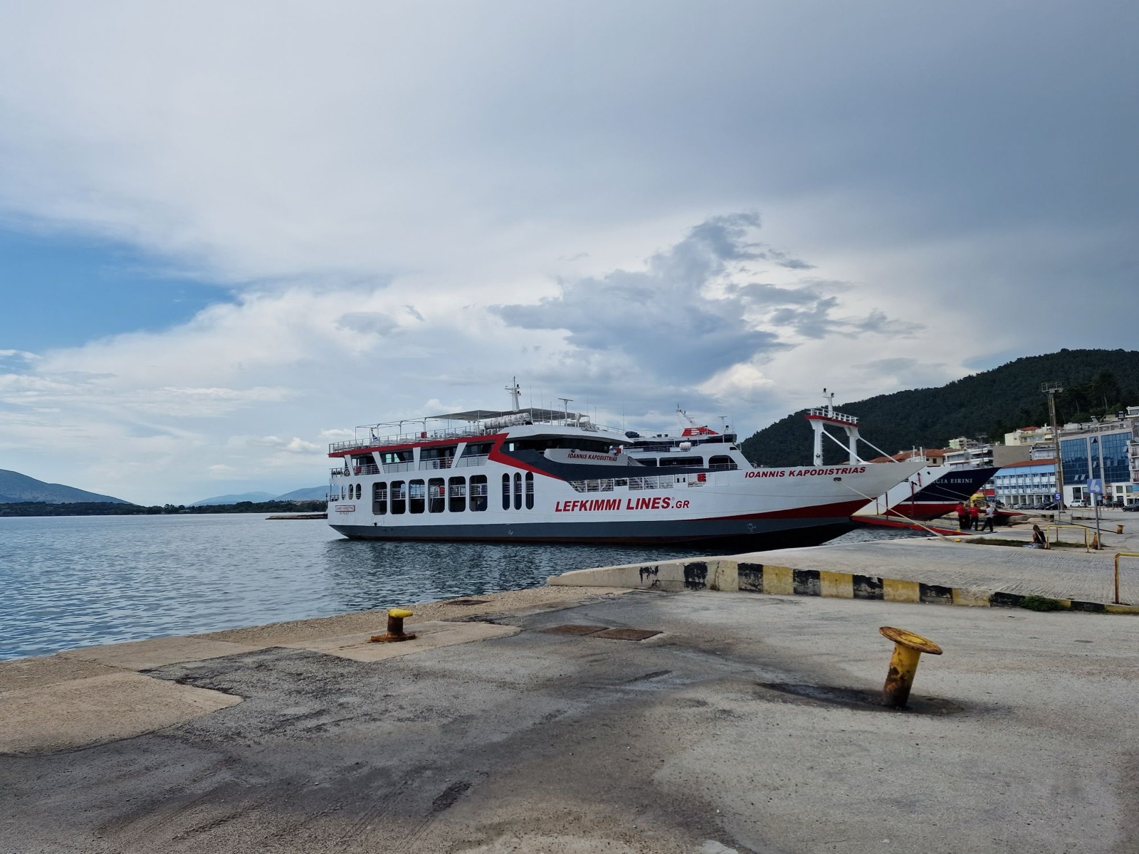 Ioannis Kapodistrias, a white car ferry with a red stripe, a dark grey lower hull and the words Lefkimmi Lines.gr along her side, moored at a dock in Igoumenitsa with a dark-hulled ferry beyond her bow.