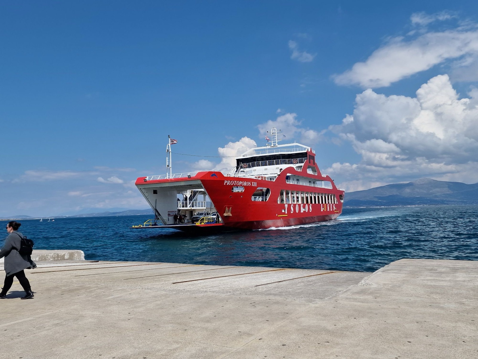 Protoporos XIII, a red Tsokos Lines double-ended car ferry, approaching the dock with her bow ramp lowered and cars and a motorbike on the open deck, hills across the water behind.