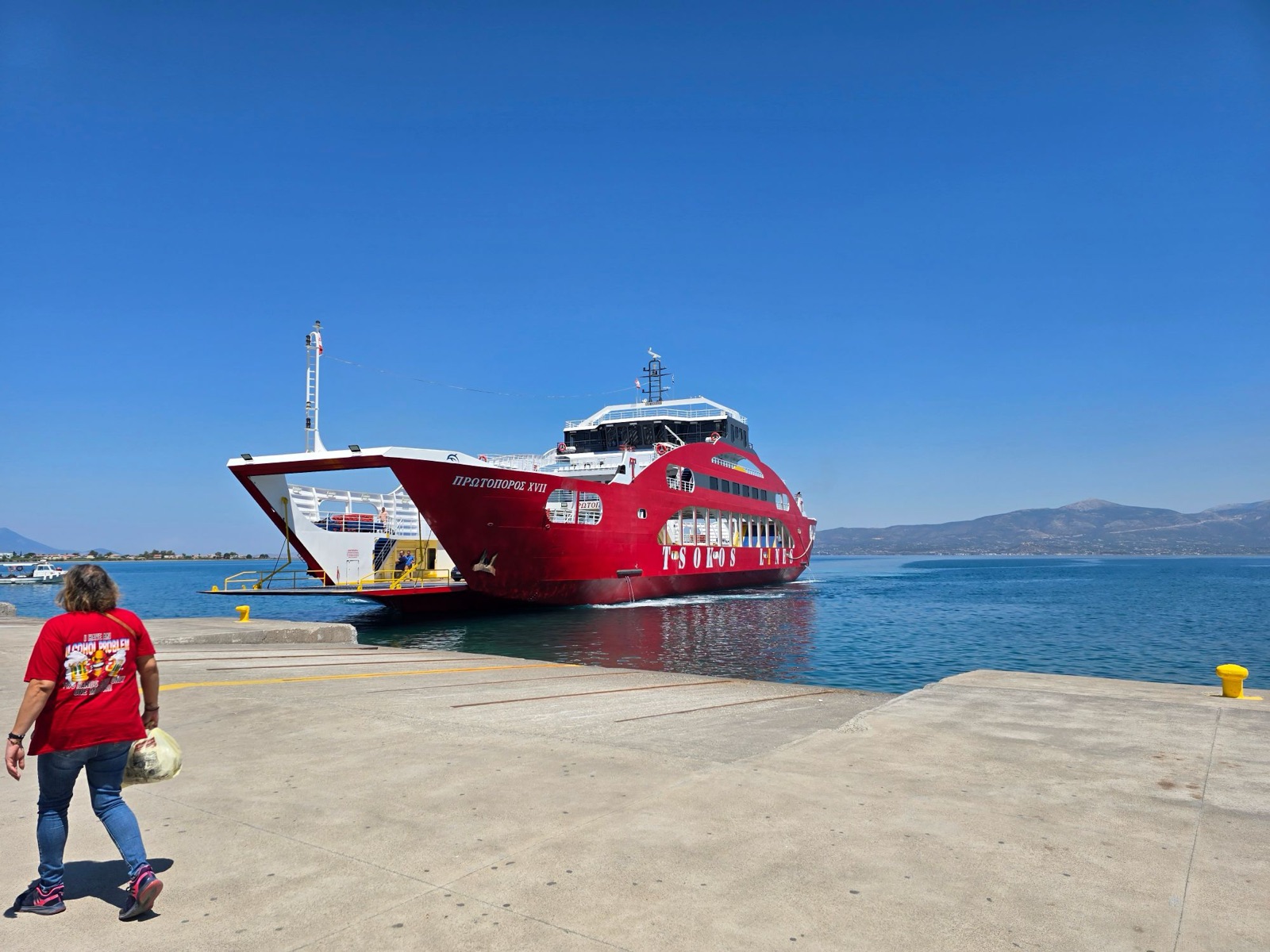 Protoporos XVII, a red Tsokos Lines double-ended car ferry with her name in Greek on the bow, arriving at a dock with the open bow ramp lowered on a clear day, hills across the water behind.
