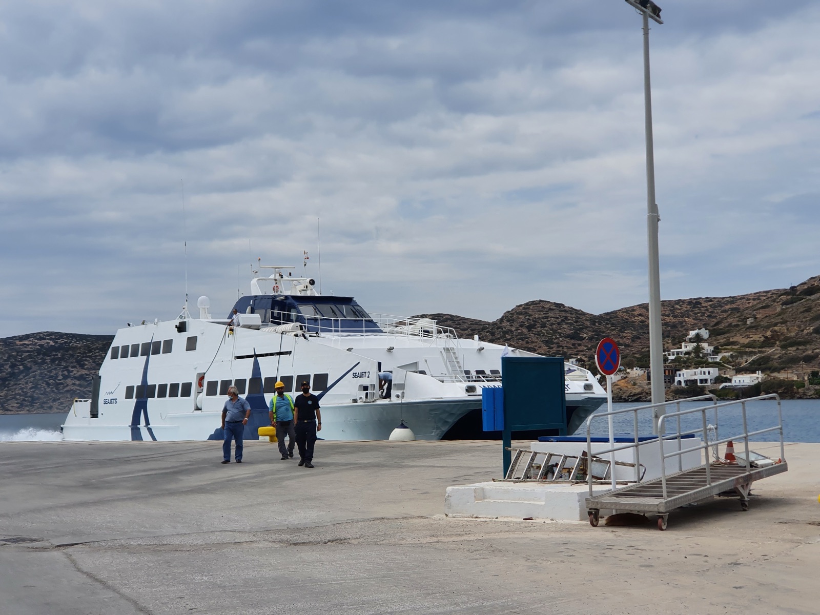 The Seajets catamaran moored at an island port in May 2021, when she still carried her earlier name Seajet 2 on her white and blue hull, with port staff walking along the dock and dry hills behind.