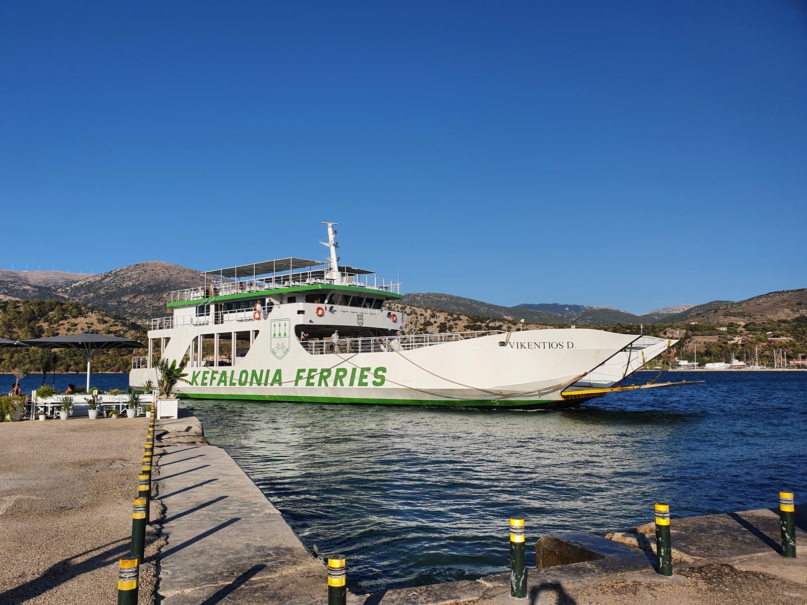 Vikentios D., a white car ferry with a green stripe and KEFALONIA FERRIES in large green letters along the hull, arriving at a small dock with her ramp lowered, an open upper deck under a canopy, and green and yellow bollards in the foreground, with hills behind.